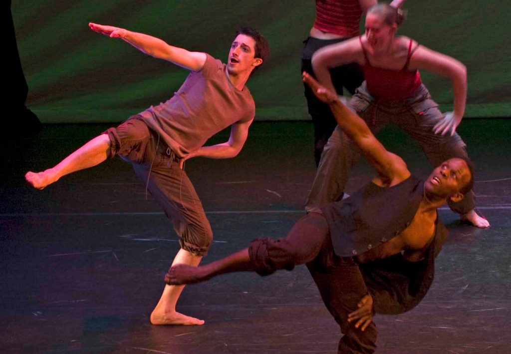 Kaleidoscope Dance Theatre: A group of dancers on stage, under pink lighting. One of their arms is extended like they’re pointing to the sky.