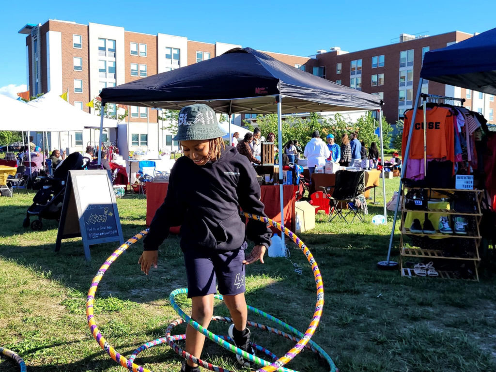 Black Artist Collective: A child is in the middle of hula hooping three hula hoops at one time - two of which have fallen to their feet. Behind them you can see a festival going on. 