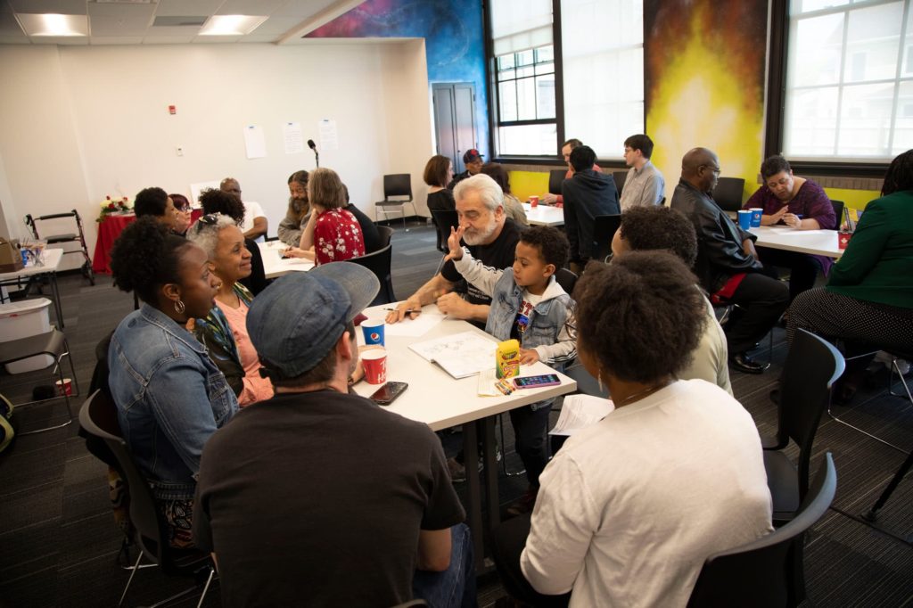 Frontline Buffalo: People are gathered around a table, in the middle of a conversation. A young child sitting at the table is raising their hand, and the person across from them is smiling at them.