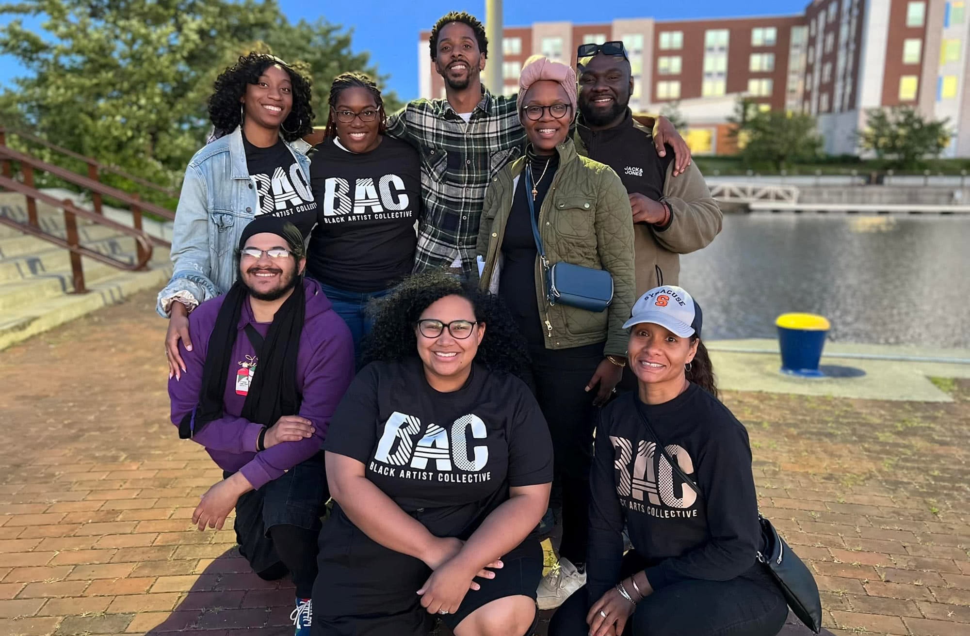 Black Artist Collective: A group of Black people with their arms wrapped around each other, looking into the camera, smiling. Some of them are wearing matching Black Artist Collective t-shirts.