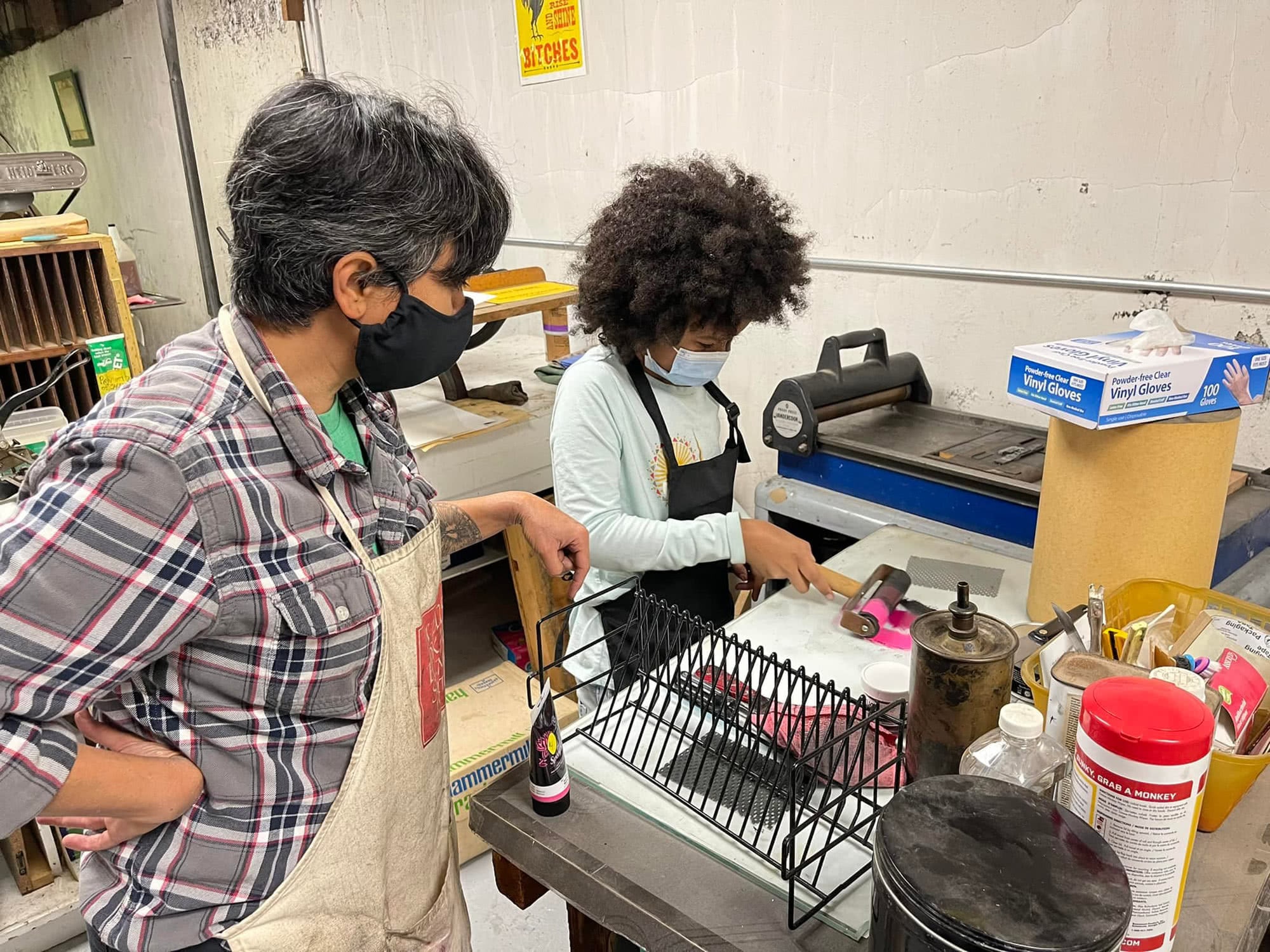 Black Artist Collective: Two people are gathered around a table in a letterpress studio, wearing aprons. One student is rolling out paint on the table to prepare for a print, and the other person is watching over them.