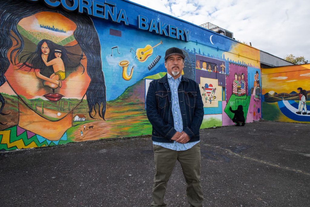 Teatro Yerbabruja: A person standing in front of a vibrant painted mural on the side of a bakery that includes images of musical instruments, landscapes, and people.