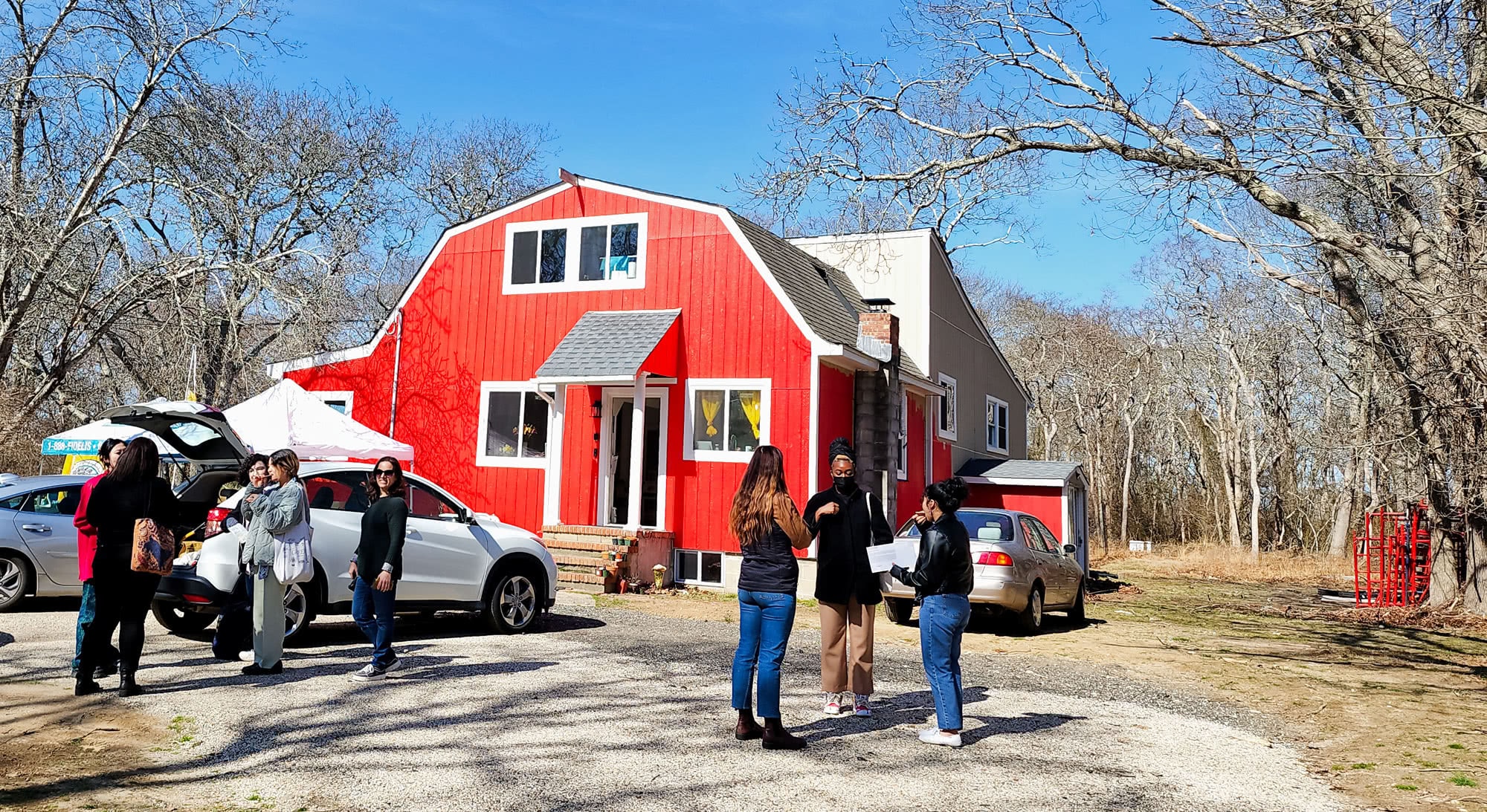 Ma’s House: A barn shaped house, painted red, against a blue sky in the background. People are gathered in small groups around the house, in mid-conversation.