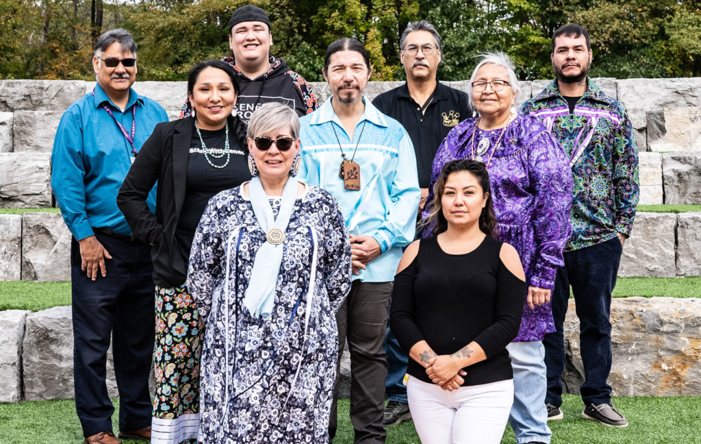 Seneca Museum: The staff of Seneca Museum, standing outside + looking into the camera. People are wearing clothes with different patterns, beaded jewelry, and Der. Joe Atahlman in the center is wearing a wooden necklace with a version of the museum logo on it - a flower sprig.
