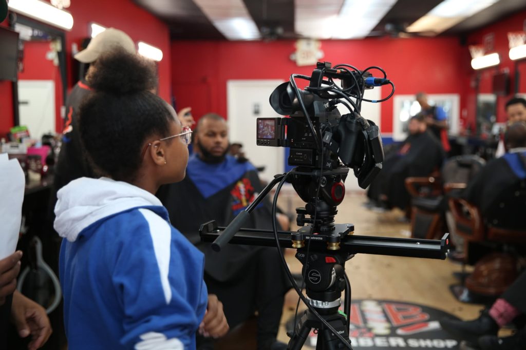 Youth FX: A youth is standing behind a camera setup. They’re set up in a barber shop, and through the viewfinder of the camera you can see that the camera frame being set up shows a person getting a haircut.