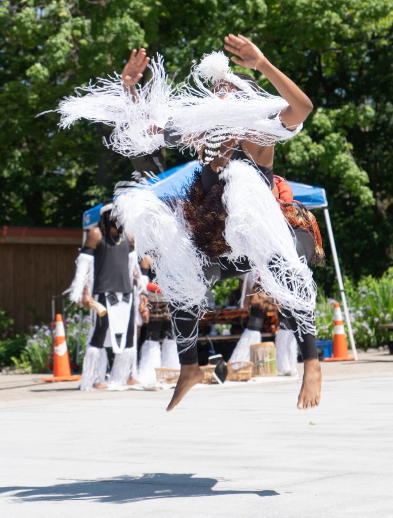 TRANSART: A dancer is jumping in the air - the fringe on their costume - around their ankles and arms - looks like it’s suspended in the air, covering their face. This is taking place at a festival that explores an afrofuturist version of Pinkser, a celebration of when enslaved Africans were freed in 17th Century NY.