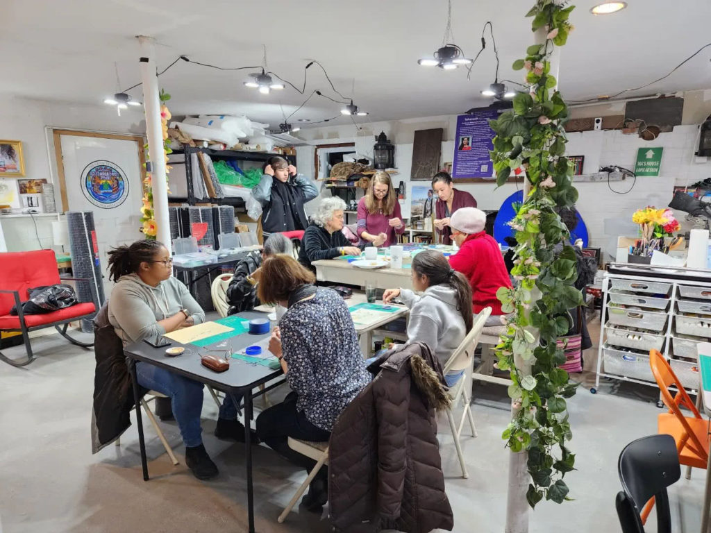 Ma’s House: A room of people gathered around tables with pliers, beads, wire, cutting mats, and other beading materials for a beading worksop. People are in mid conversion, or are looking at others to see what they’re doing.