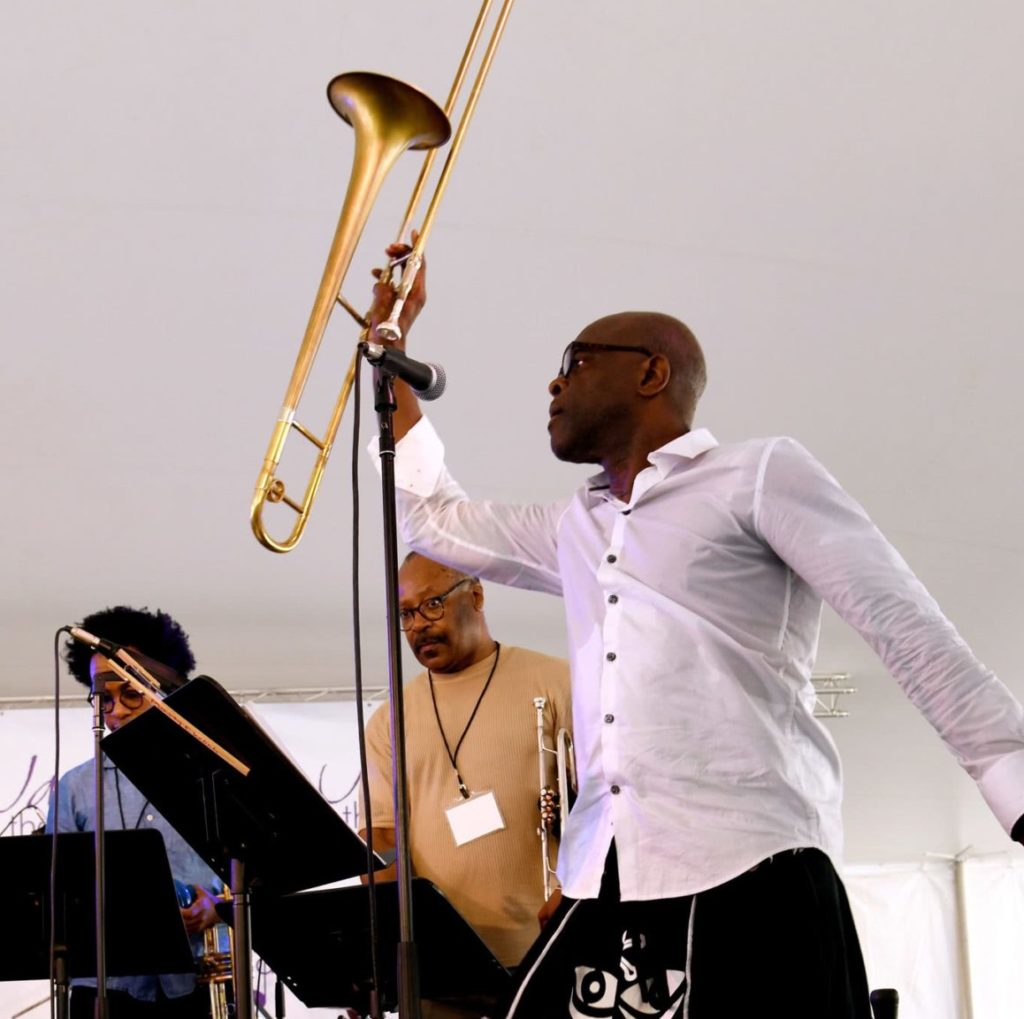 TRANSART: A trombone player standing in front of a mic triumphantly holds their trombone up in the air. Behind them are other musicians - a trumpet and drummer - reading music from the music stands in front of them.