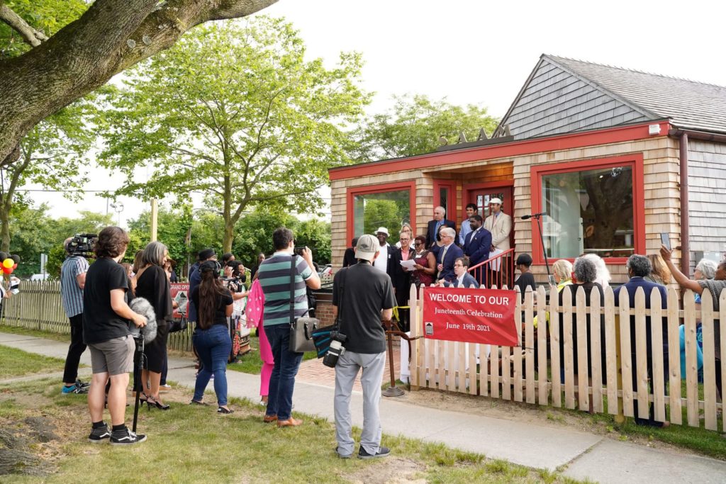 Southampton African American Museum: A group of people stand on the steps of a building that has brown shingles and red trim around the windows and doors. In front of the building on the fence is a red sign that reads “Welcome to Our Juneteenth Celebration”. A small crowd is gathered, looking on and and taking pictures/videos.
