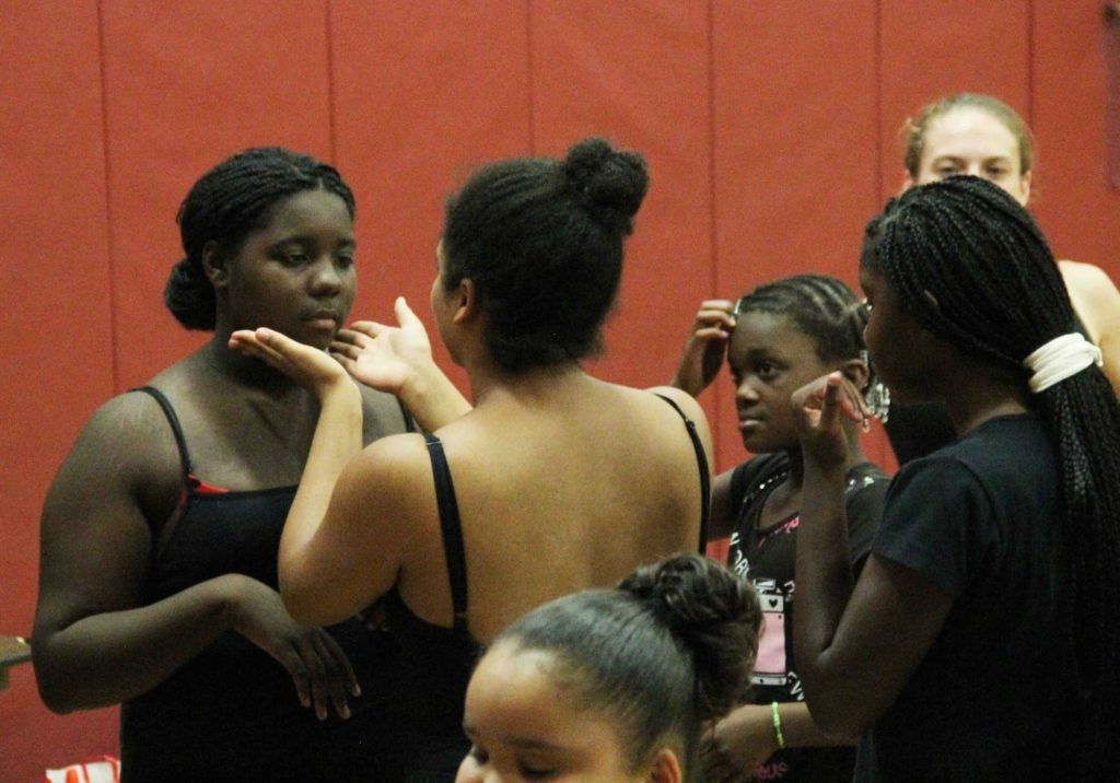 Kaleidoscope Dance Theatre: A dance teacher holds a students’ face cupped in her hands. The student looks a bit sad and appears to be looking down at the ground, and the dance teacher is lifting their head up.