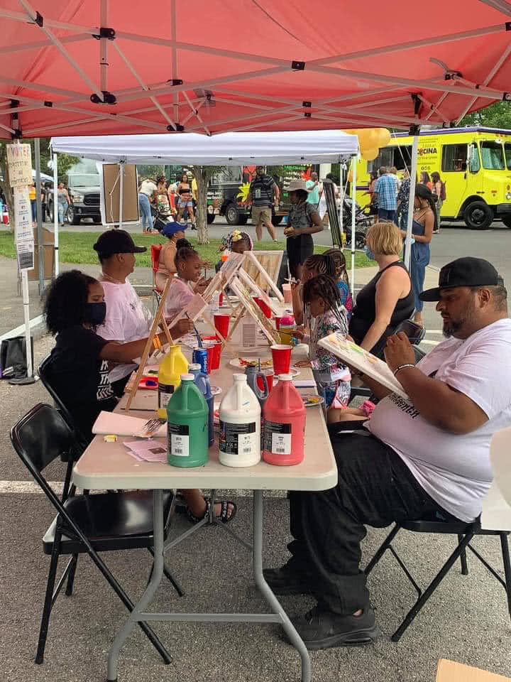 Black Artist Collective: A group of people - adults and children - sit on either side of a long table under a tent. On the table there are gallons of paint, easels and canvases, paintbrushes, and other art materials - people are painting. Behind them you can see the rest of a festival taking place.